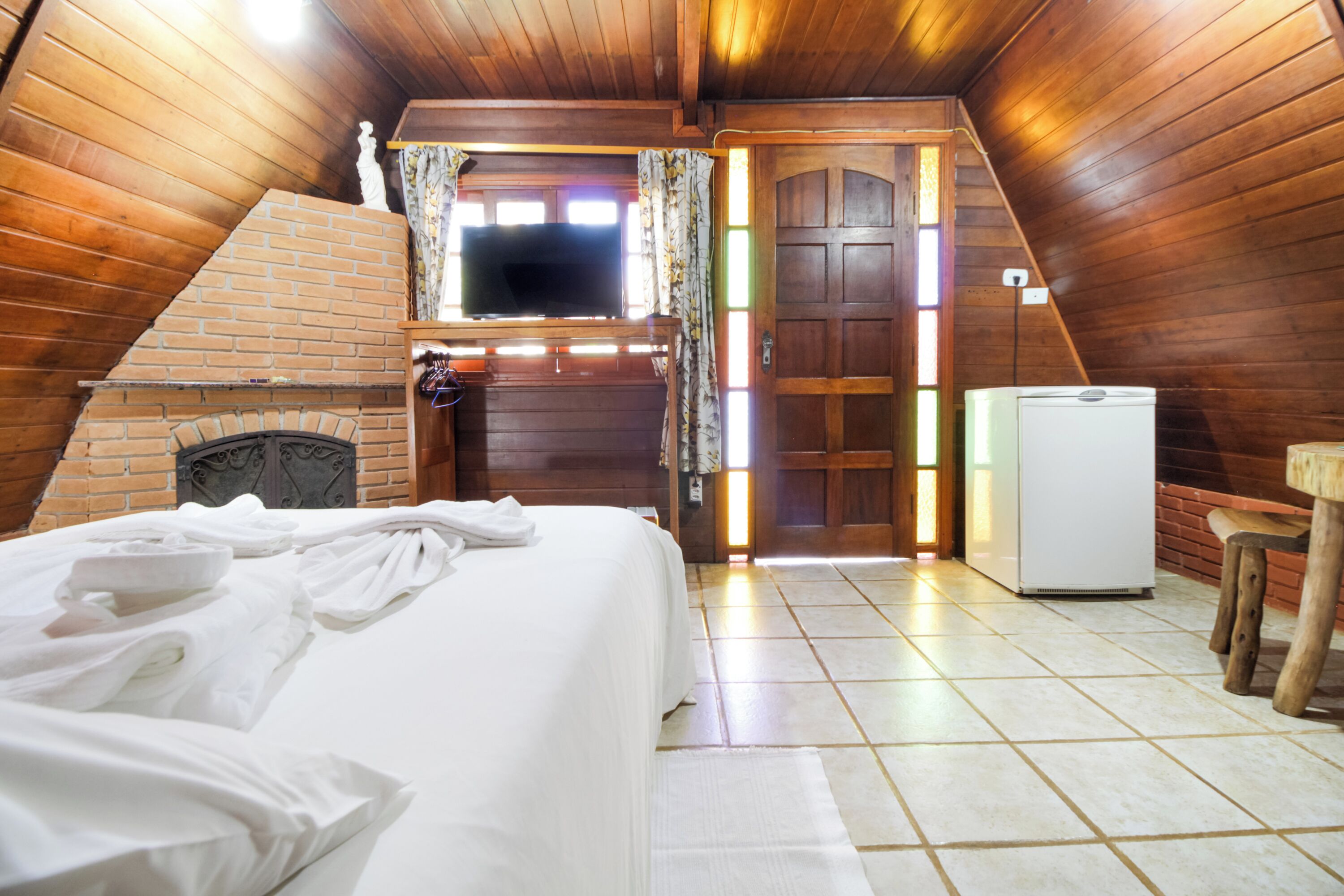 Un dormitorio acogedor en el ático con chimenea de ladrillo, una cama con ropa blanca, un televisor y un refrigerador.
