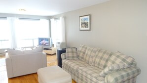 Una sala de estar con un sofá blanco, un sofá a cuadros, un otoman blanco, un televisor y un cuadro en la pared.