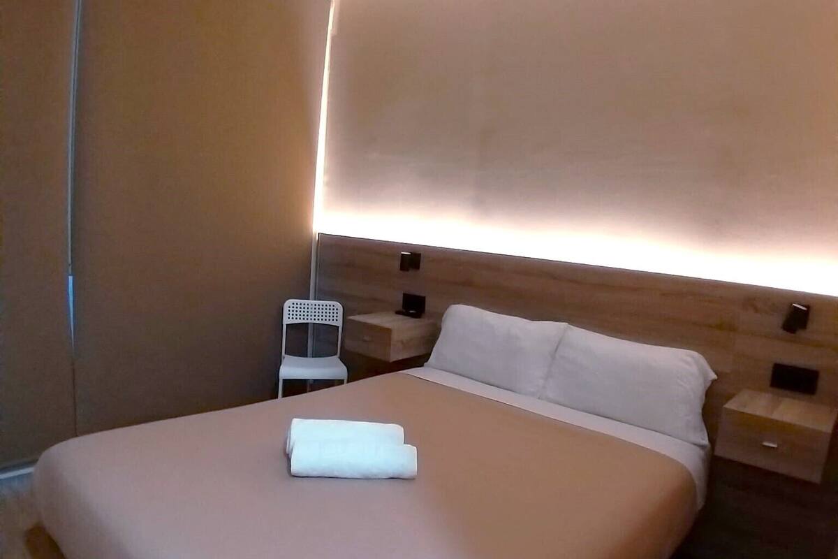 Une chambre d’hôtel moderne avec un grand lit, une table de chevet, une petite table et une lampe fixée au mur.