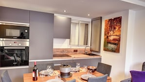 A modern kitchen with a Bosch oven, a dining area with a wooden table set for four, and a large window with blinds. - Cozy apartment between lake and mountains (Annecy)