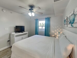 Une chambre à coucher avec un lit, un téléviseur posé sur un meuble blanc, un ventilateur de plafond, une fenêtre avec des rideaux et un mur orné d’un tableau représentant une tortue.