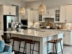 A modern kitchen with white cabinetry, a large island with bar stools, a stainless steel refrigerator, and a hanging light fixture.