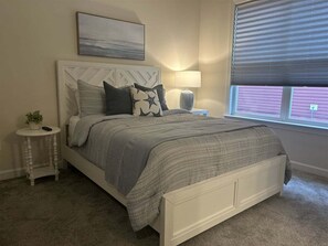 Ein Schlafzimmer mit einem Bett, einem Nachttisch, einer Lampe, einem Fenster mit Jalousien und einem Bild an der Wand.