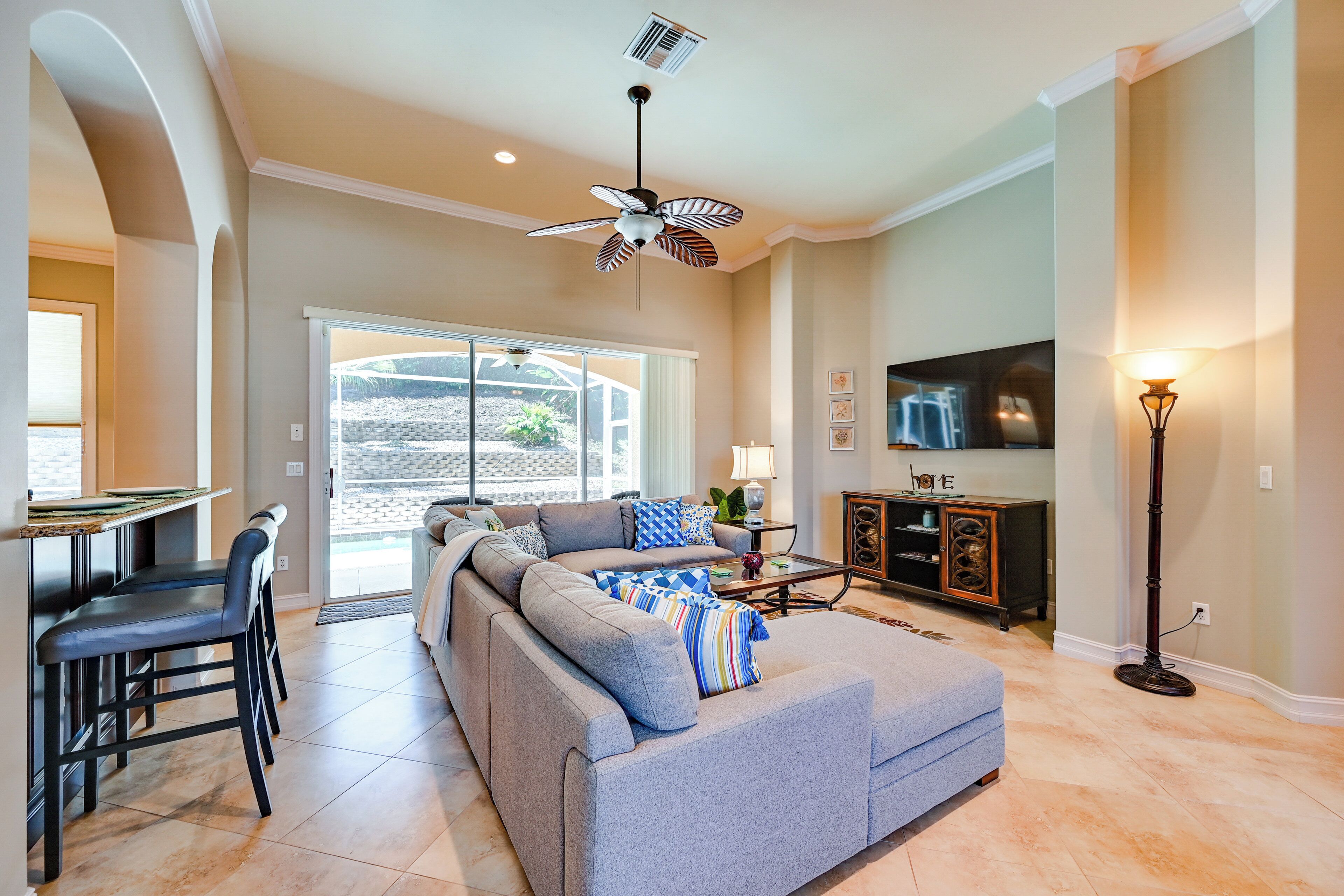 A spacious living room with a large grey sectional sofa, a dining area with bar stools, a television, and a ceiling fan.