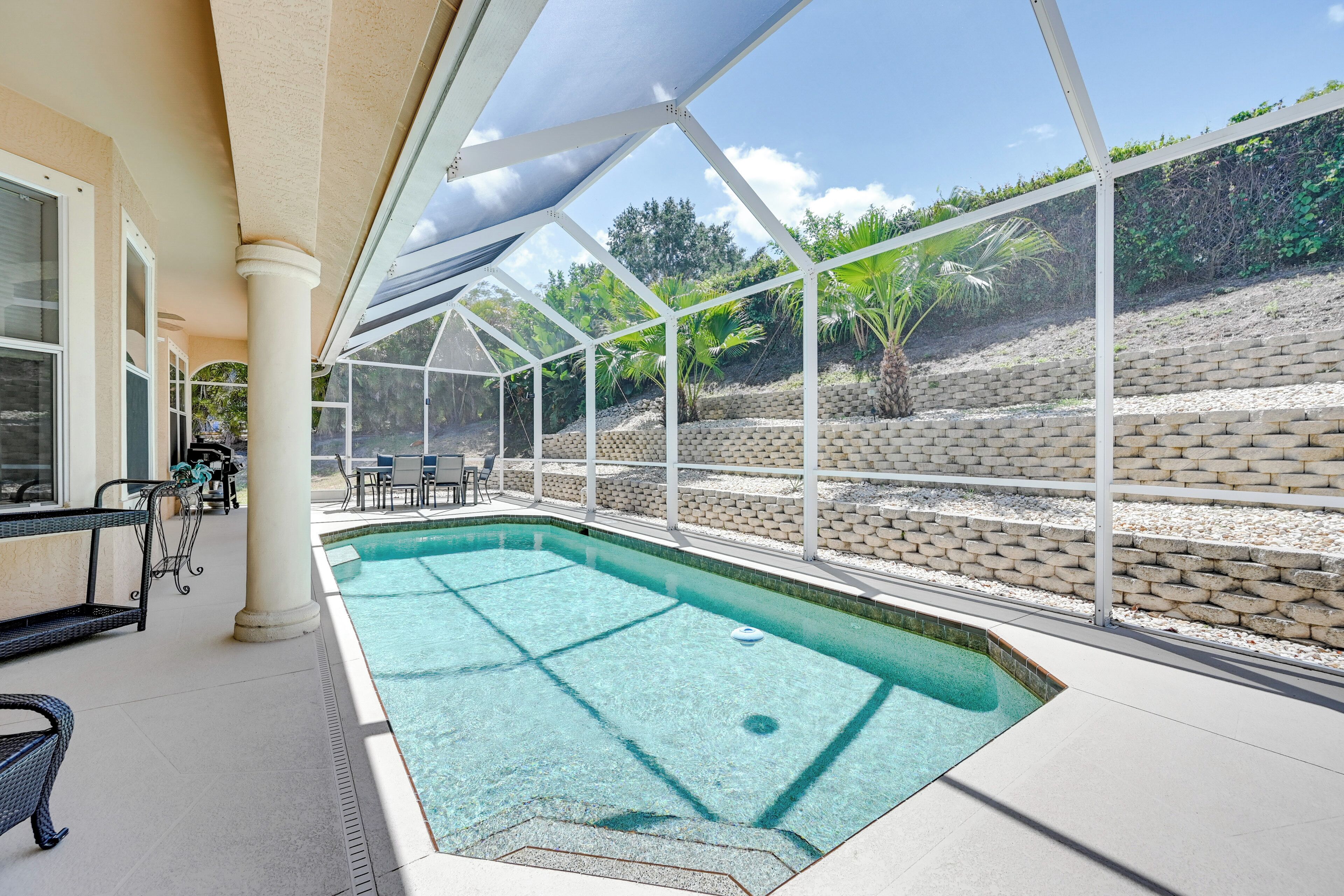 An indoor pool with clear blue water, surrounded by a covered patio area with seating and a view of a landscaped garden.