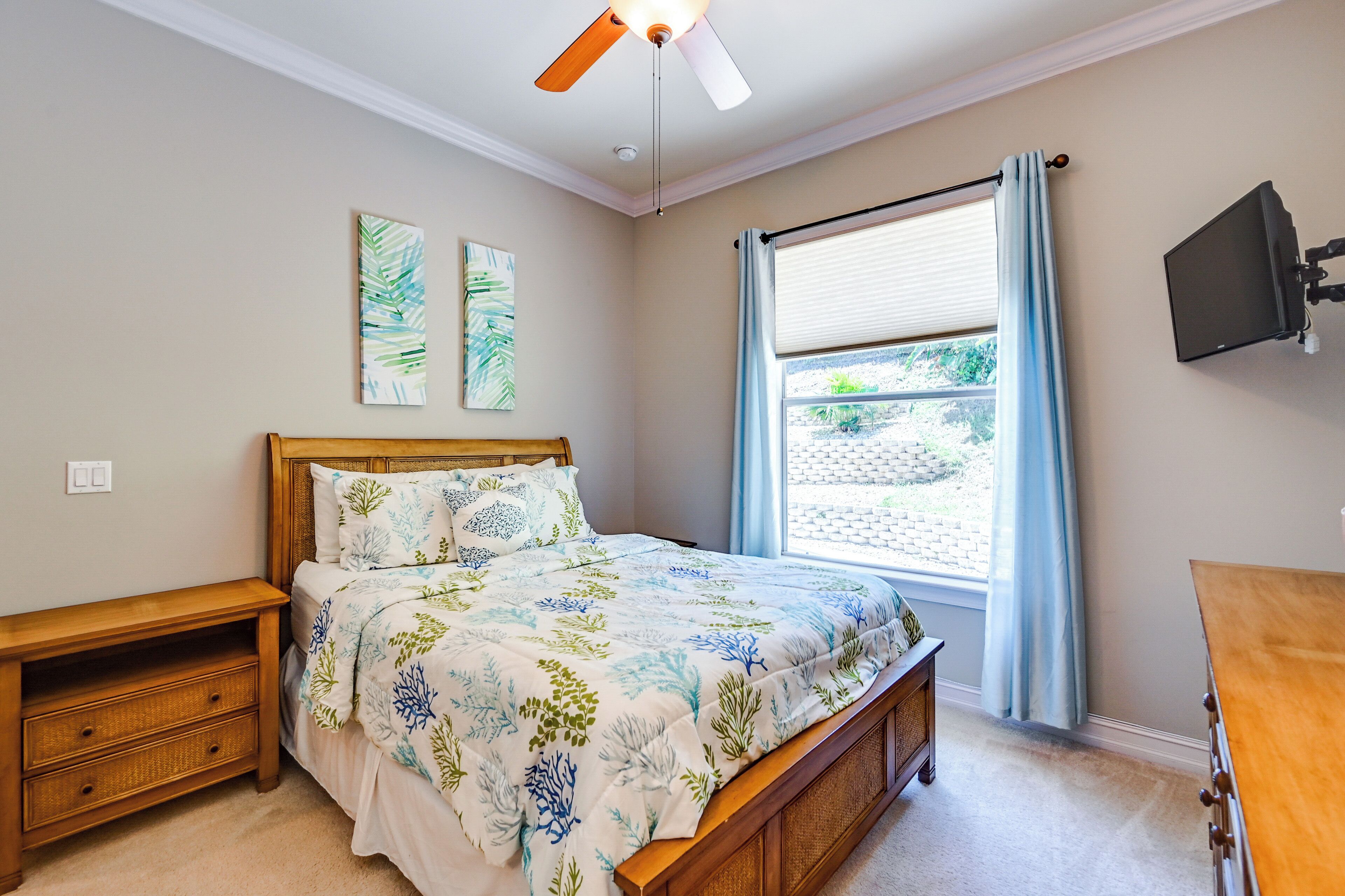 A bedroom with a bed, wooden nightstand, ceiling fan, artwork, window with curtains, and a TV mounted on the wall.