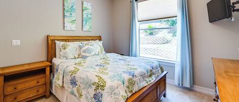 A bedroom with a bed, wooden nightstand, ceiling fan, artwork, window with curtains, and a TV mounted on the wall.