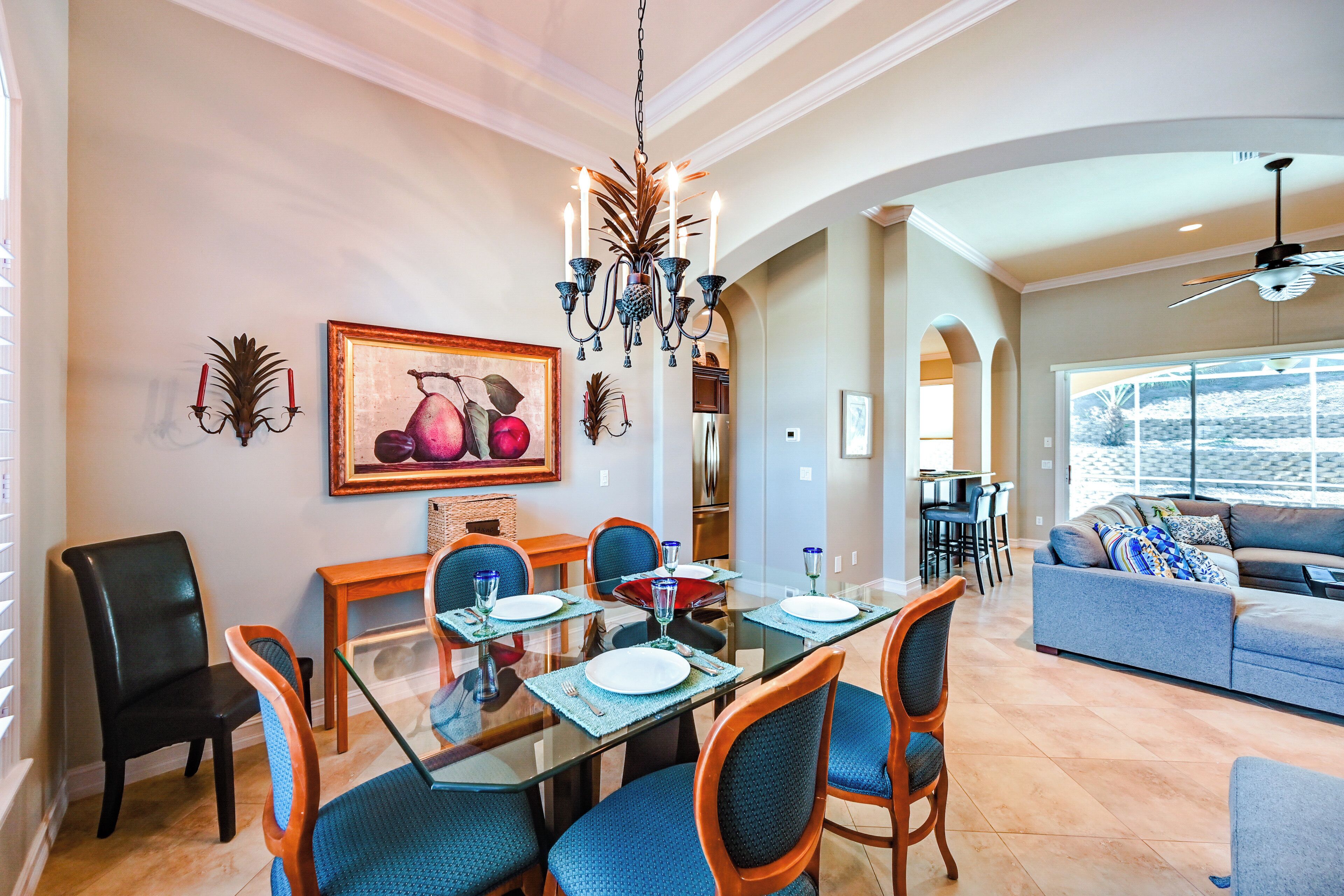 A dining area with a glass table, blue chairs, and a chandelier. There is a painting on the wall and a sofa in the background.
