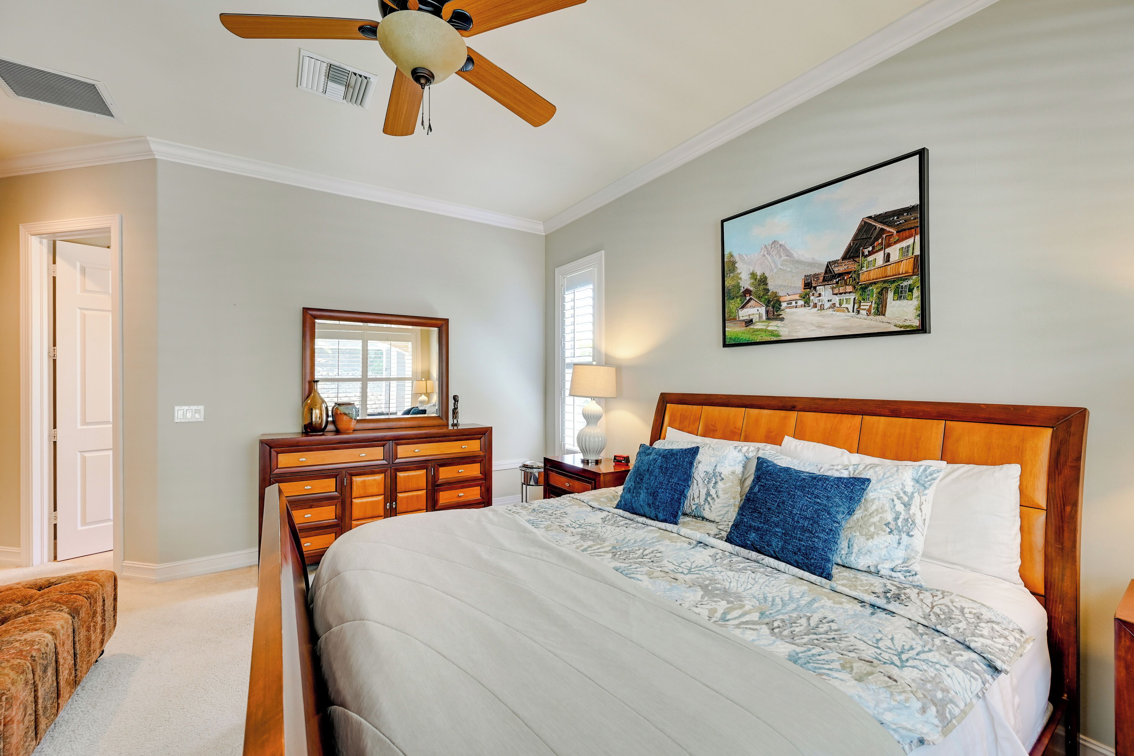 A bedroom with a large bed, wooden headboard, a dresser, a ceiling fan, a painting, and a window with blinds.