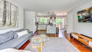 Una sala moderna con un sofá gris, una mesa de centro de vidrio y un cuadro grande de árboles. Se ve una cocina con armarios blancos y una zona de comedor con sillas blancas al fondo.