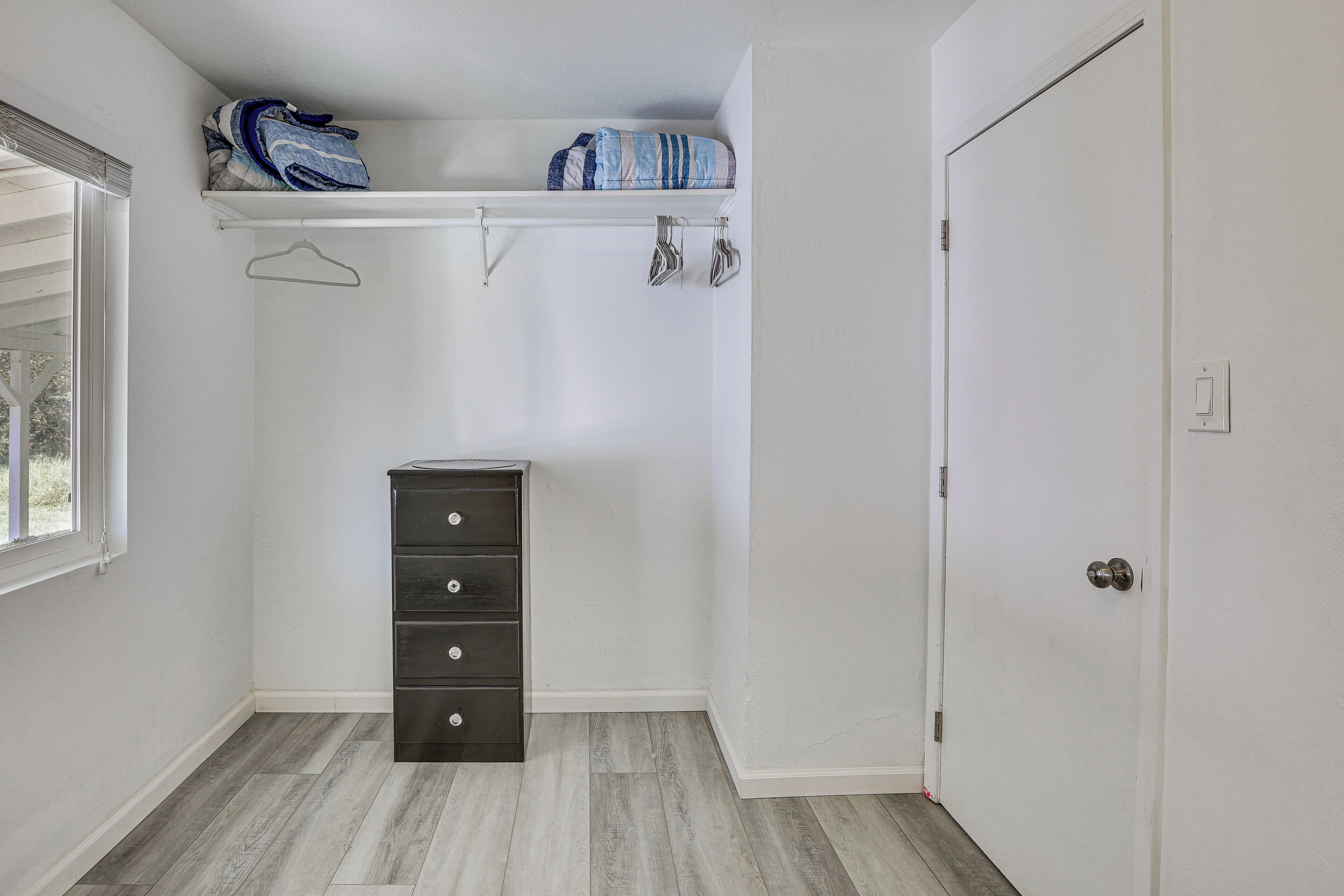 A small, well-lit room with a wooden floor, a dark-colored dresser, a white door, and a window with blinds.