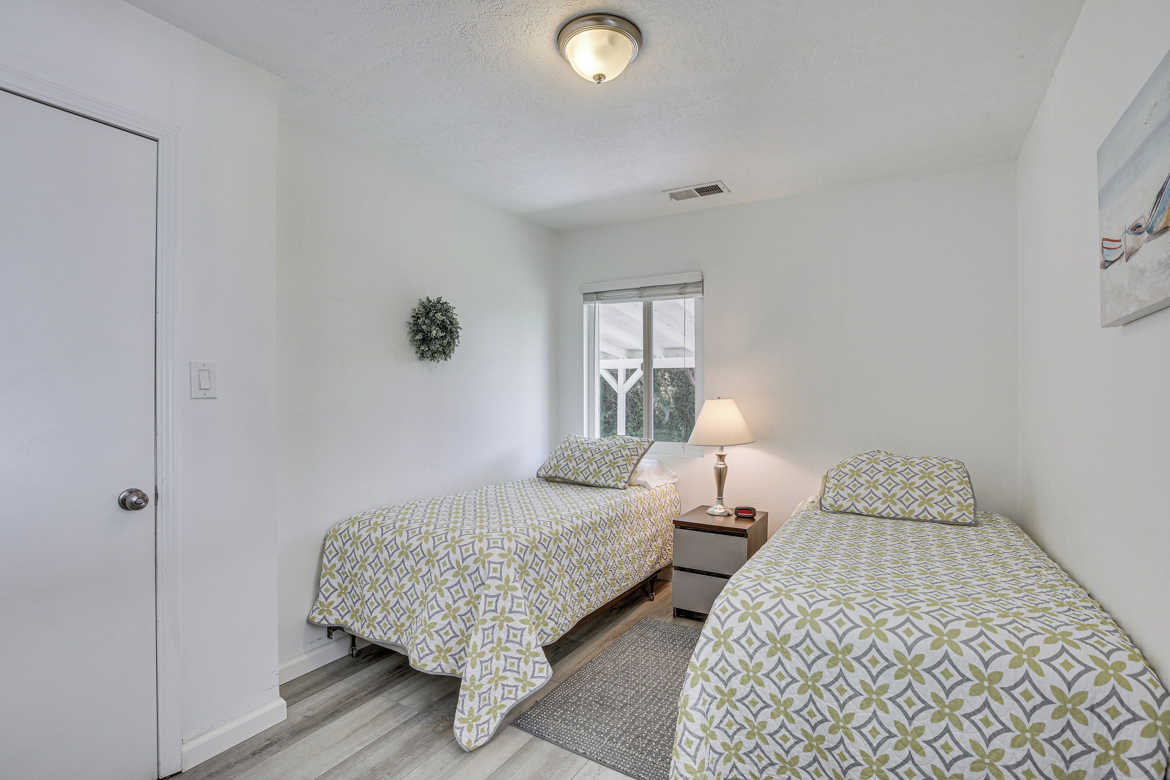 A bedroom with two single beds, a nightstand, a lamp, a window, and a door.