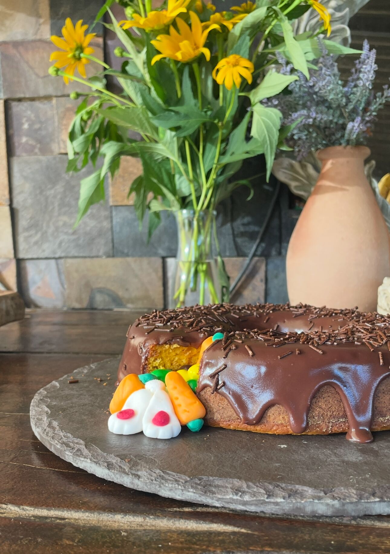 A chocolate cake with sprinkles and colorful decorations on a stone plate, with a vase of flowers in the background.