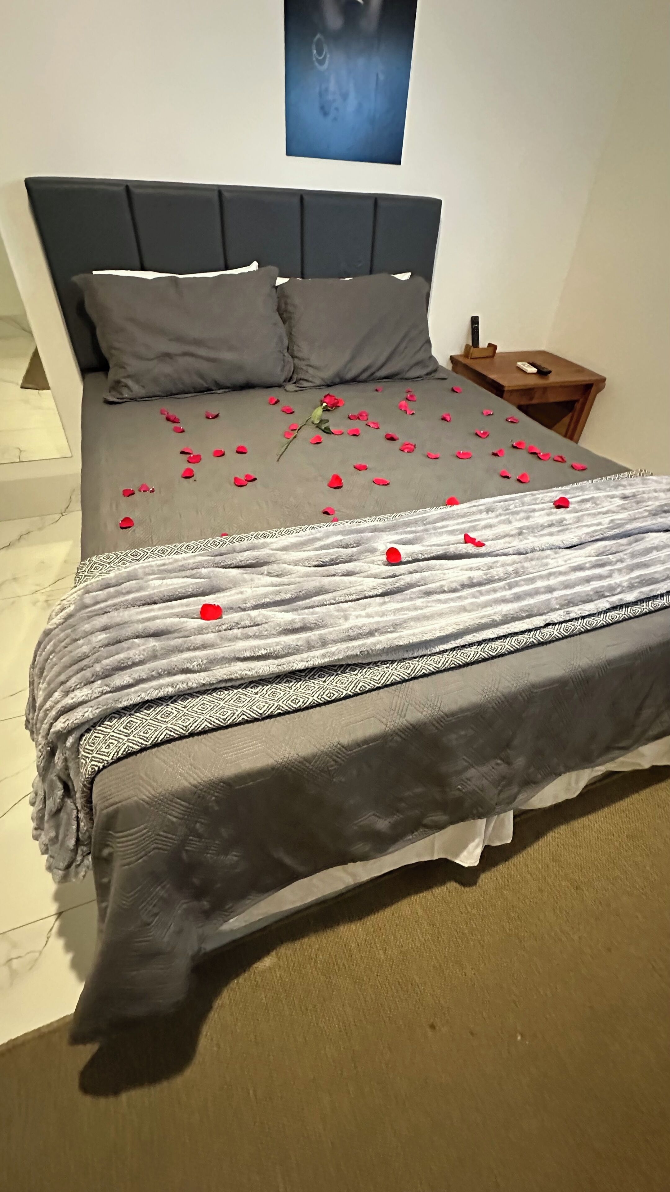 Ein ordentlich bezogenes Bett mit grauem Kopfteil, einer gemusterten Tagesdecke und roten Rosenblättern, die auf dem Bett verteilt sind.