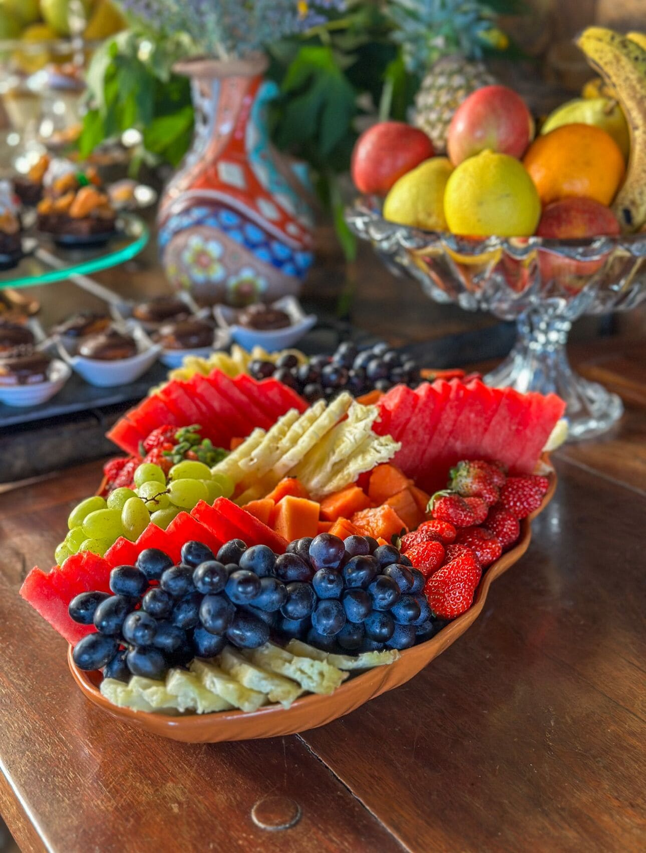 A fruit platter with various fruits and a decorative vase in the background.