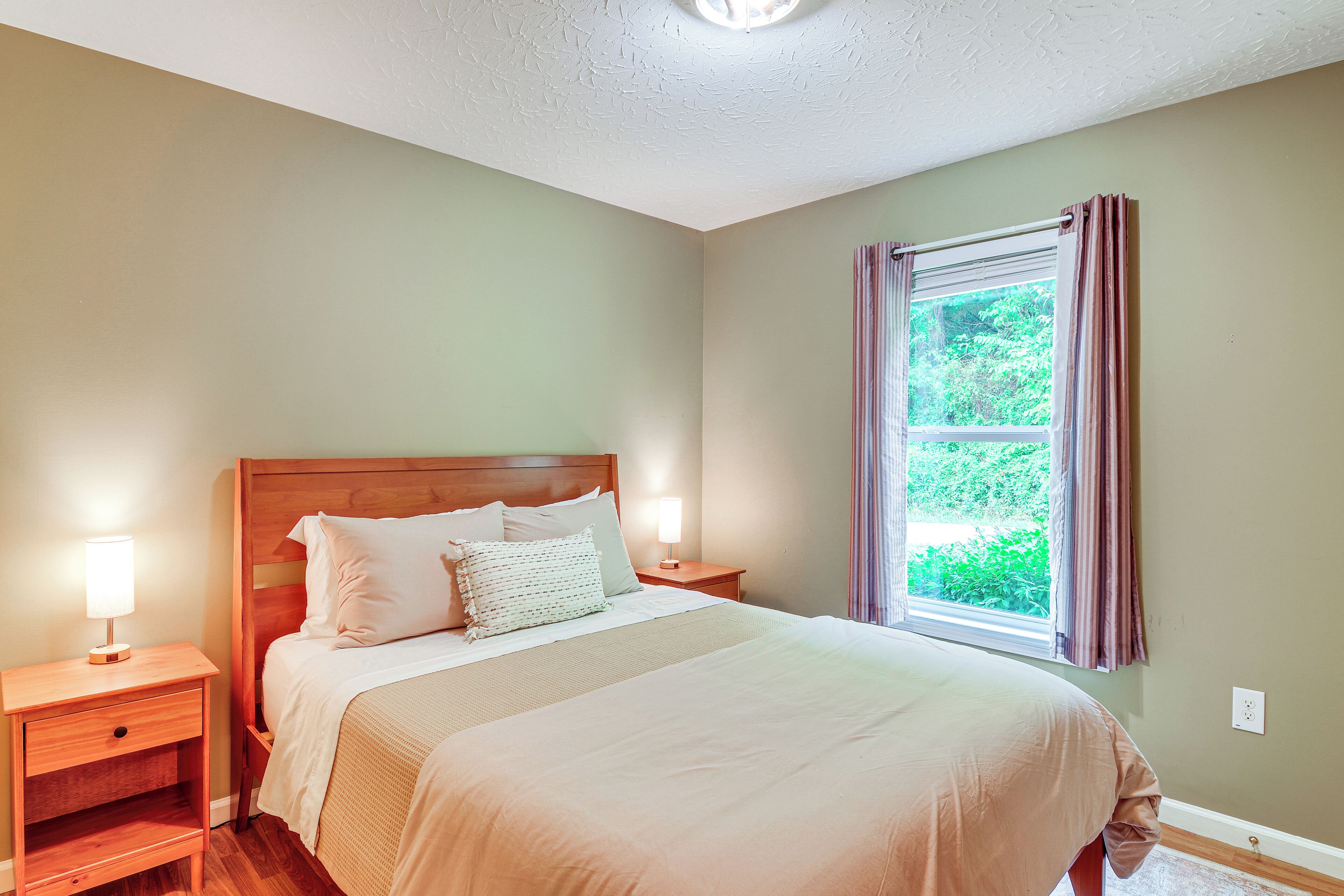 Una cama bien tendida con sábanas blancas y cabecera de madera, una mesita de noche con lámpara, y una ventana con cortinas.