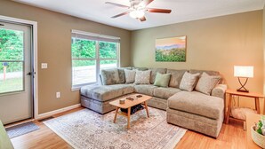 A living room with a sectional sofa, a wooden coffee table, a ceiling fan, and a large window with a view of greenery. - Campton Cottage w/ Hot Tub, Near Hiking + Climbing (Campton)