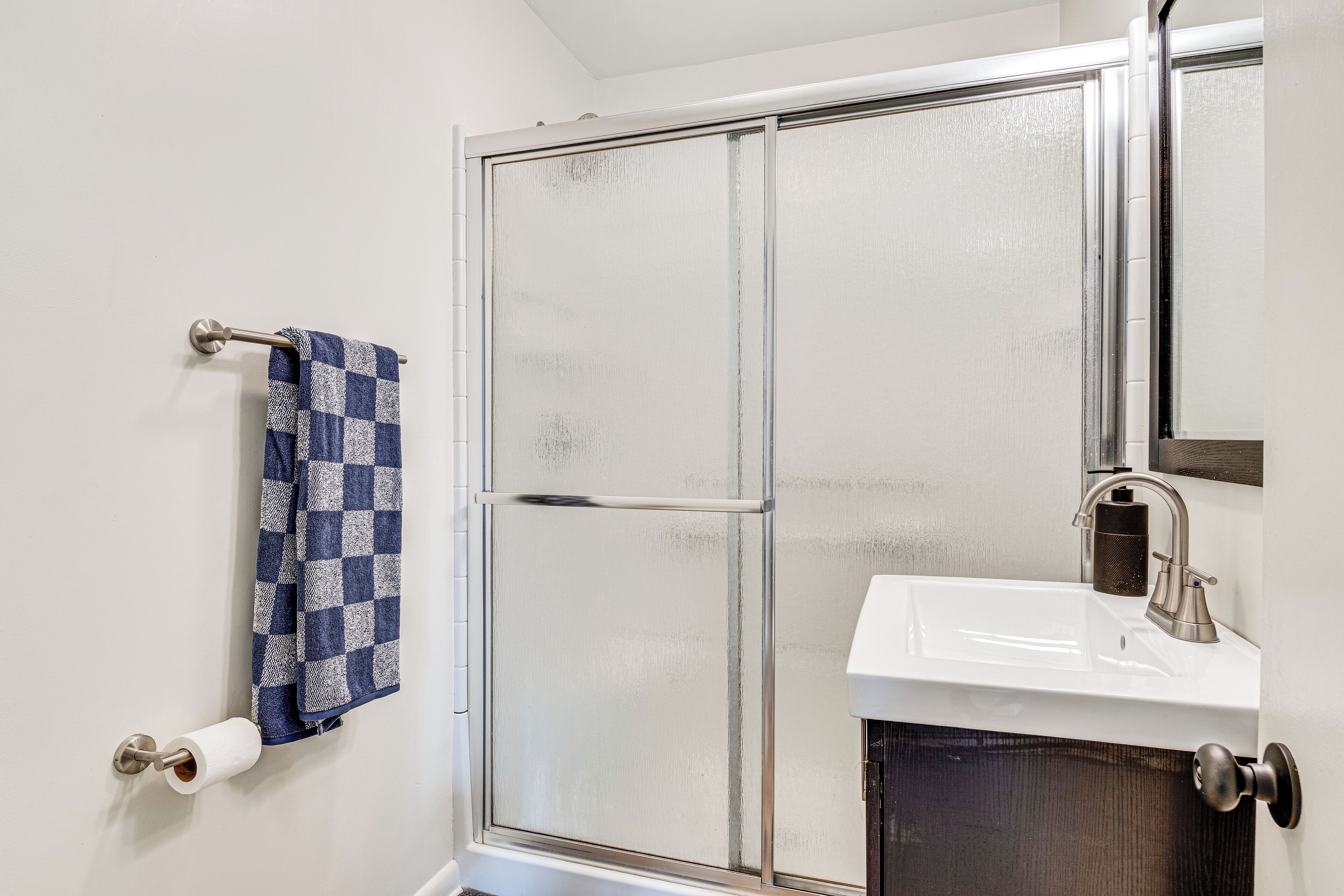 Un baño con cabina de ducha, lavamanos, toallero con una toalla a cuadros y dispensador de papel higiénico.
