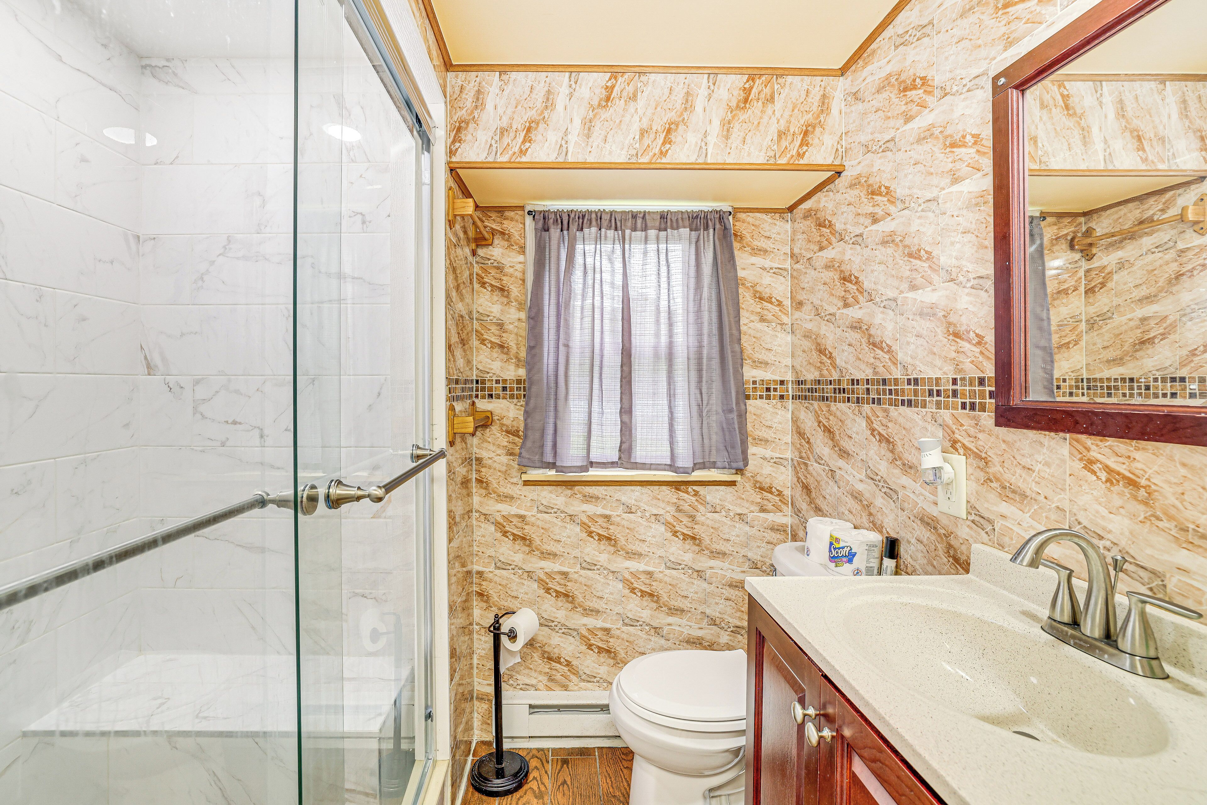Ein Badezimmer mit Duschkabine aus Glas, Toilette, Waschbecken mit Spiegel und einem Fenster mit Vorhang.