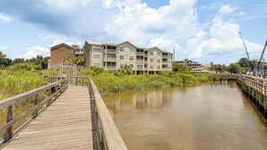 A wooden boardwalk leading to a multi-story building with balconies, situated beside a calm river.