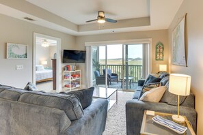 Ein Wohnzimmer mit Sofa, Couchtisch, Fernseher, Bücherregal und einem Balkon mit Sitzgelegenheit im Freien.