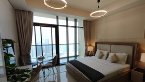 A modern hotel room with a large bed, a desk with a chair, a view of the city through large windows, and a balcony. - Lux BnB Aykon City I Skyline Canal Views (Dubai)