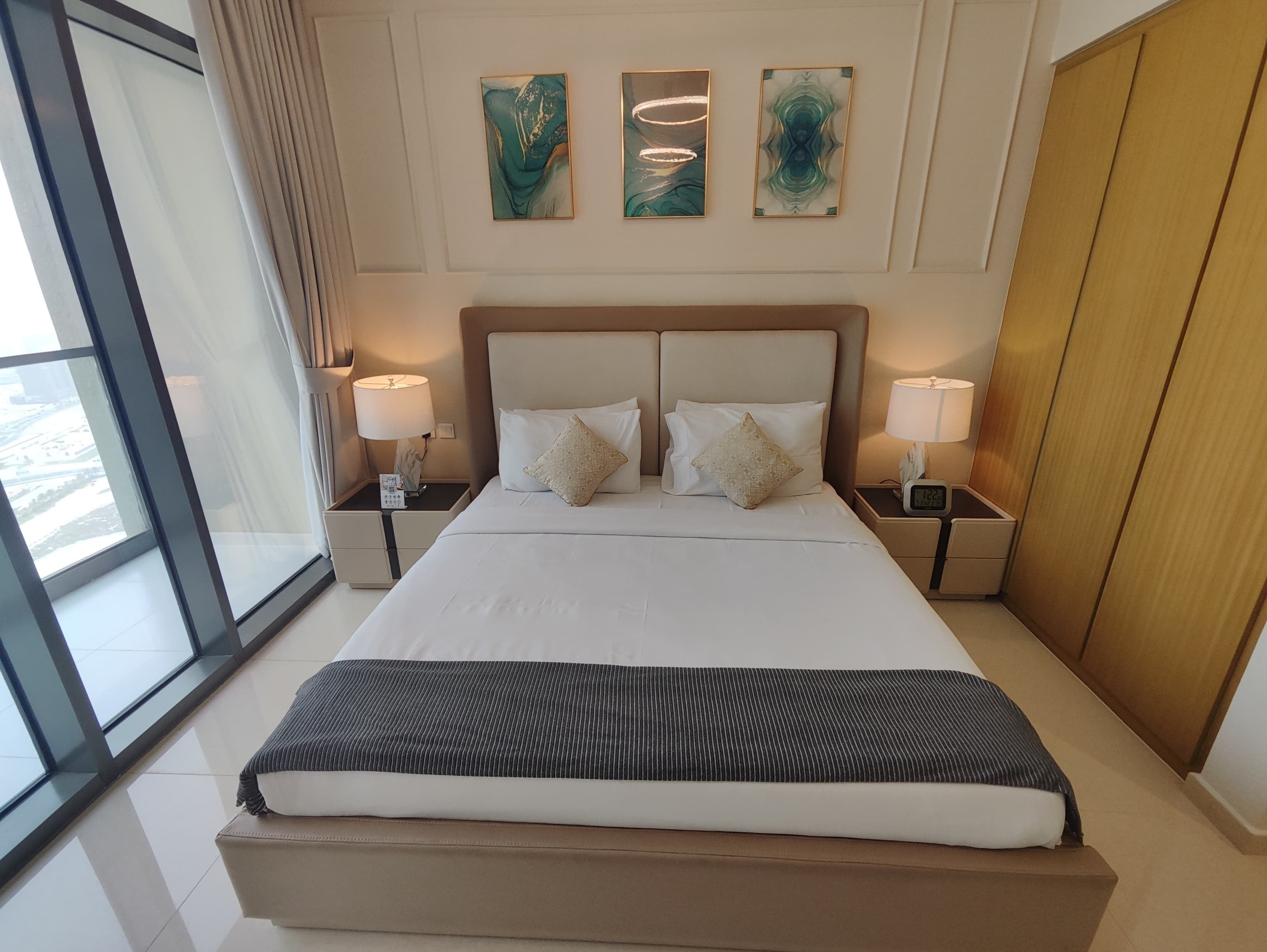 A hotel room with a large bed, two bedside tables with lamps, and three abstract paintings on the wall.