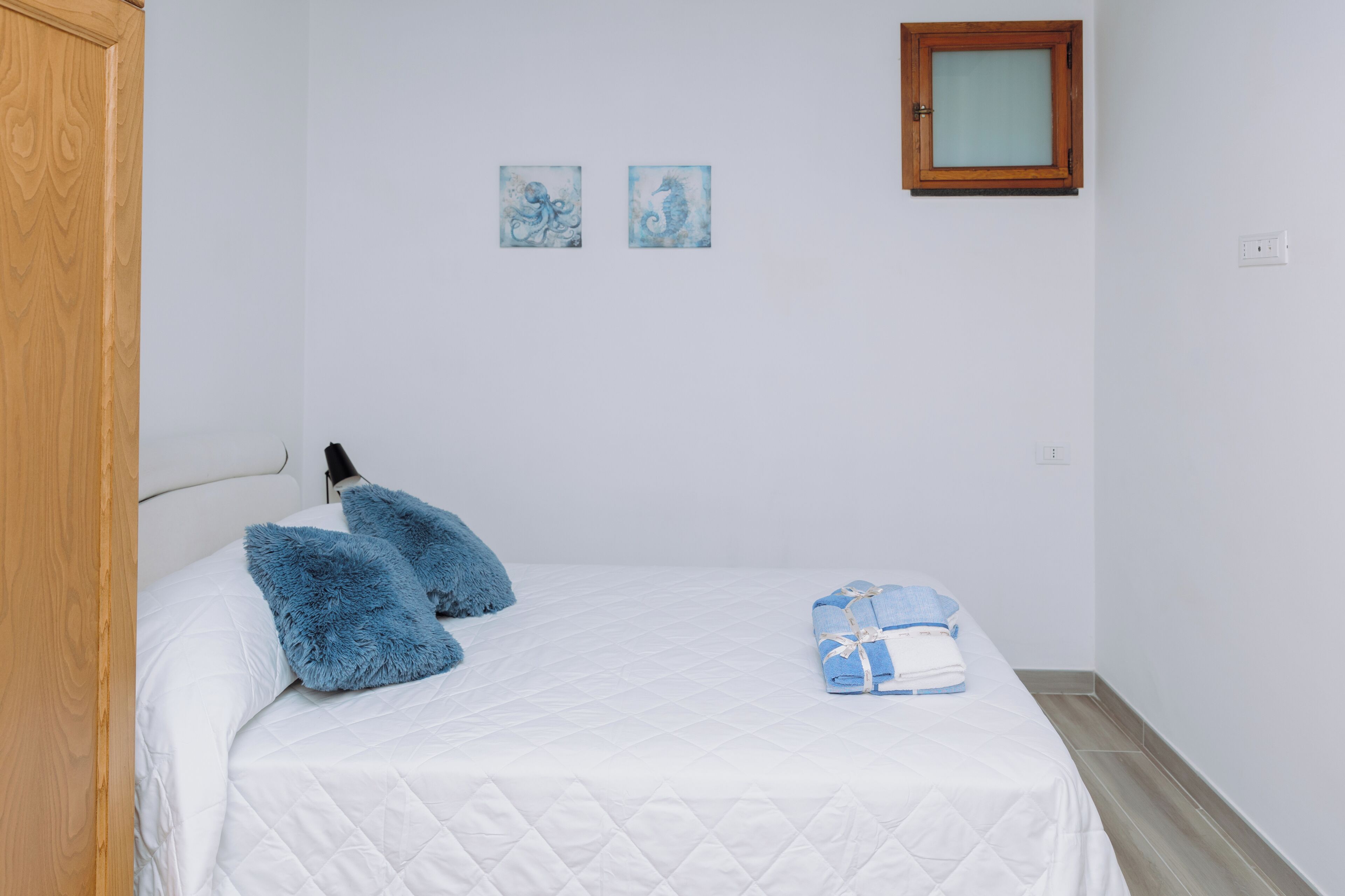 A bedroom with a white bed, two blue pillows, a folded towel, and two framed artworks on the wall.