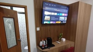 Une pièce de taille modeste, avec un bureau en bois, une chaise rouge, un téléviseur fixé sur un panneau en bois, une poubelle et un appareil de climatisation.