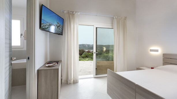 A bedroom with a large bed, a television, a bathroom, and a sliding glass door to a balcony. - HOTEL BORGO DELL'ORSO (Palau)