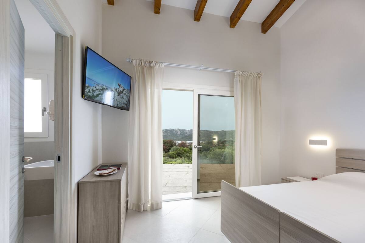 A bedroom with a large bed, a television, a bathroom, and a sliding glass door to a balcony.
