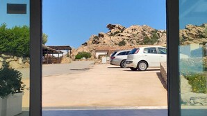 A view through a glass door of a parking area with a white car, a stone building, and a rocky hill in the background. - HOTEL BORGO DELL'ORSO (Palau)