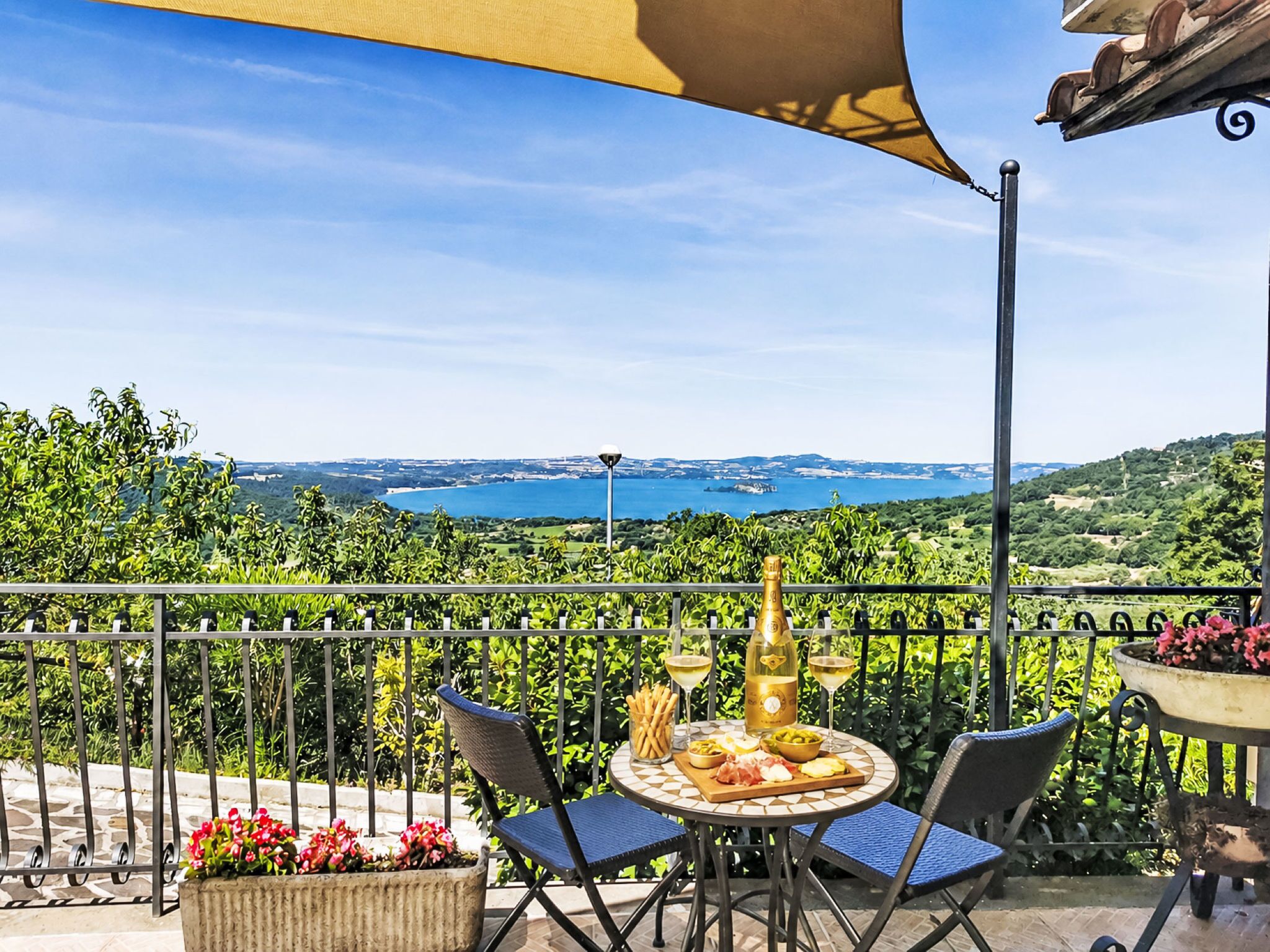 Un balcon avec une table dressée pour le dîner, une bouteille de vin et une vue sur un plan d’eau et des collines.