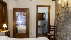 A bedroom with a wooden door, a bed, a nightstand with a lamp, and a stone wall.