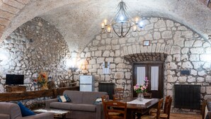 A rustic stone-walled room with a stone fireplace, a wooden dining table with chairs, a sofa, and a television on a wooden stand. - La Capraccia by Interhome (Lago di Bolsena)