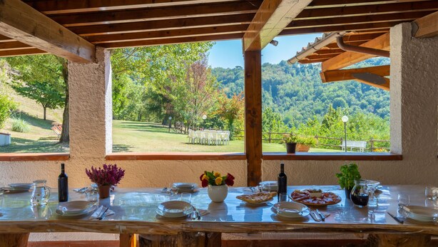 Area pranzo all'aperto con tavolo di legno apparecchiato per un pasto, con vista su un paesaggio verdeggiante tra alberi e colline.
