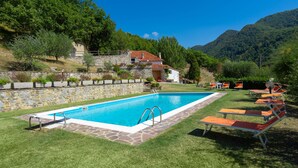 Una piscina con bordo in pietra, circondata da erba e un muro in pietra. Si notano anche sdraio e un edificio con tetto rosso sullo sfondo.