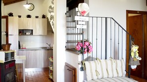 Cozinha acolhedora com fogão a lenha, pia branca e painel de azulejos. Escada com corrimão de metal leva a um patamar onde um boneco está exposto.