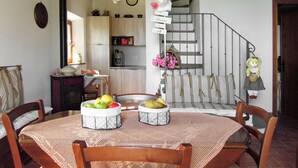 Uma área de refeições com uma mesa redonda, cadeiras e uma cesta de frutas. Há uma escada que leva a um andar superior.