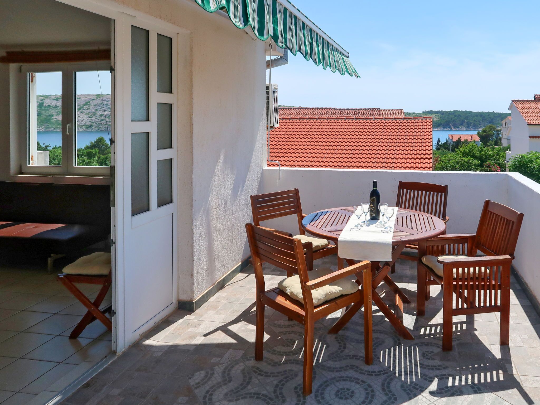 En terrasse med et rundt bord, seks træstole og en flaske vin.