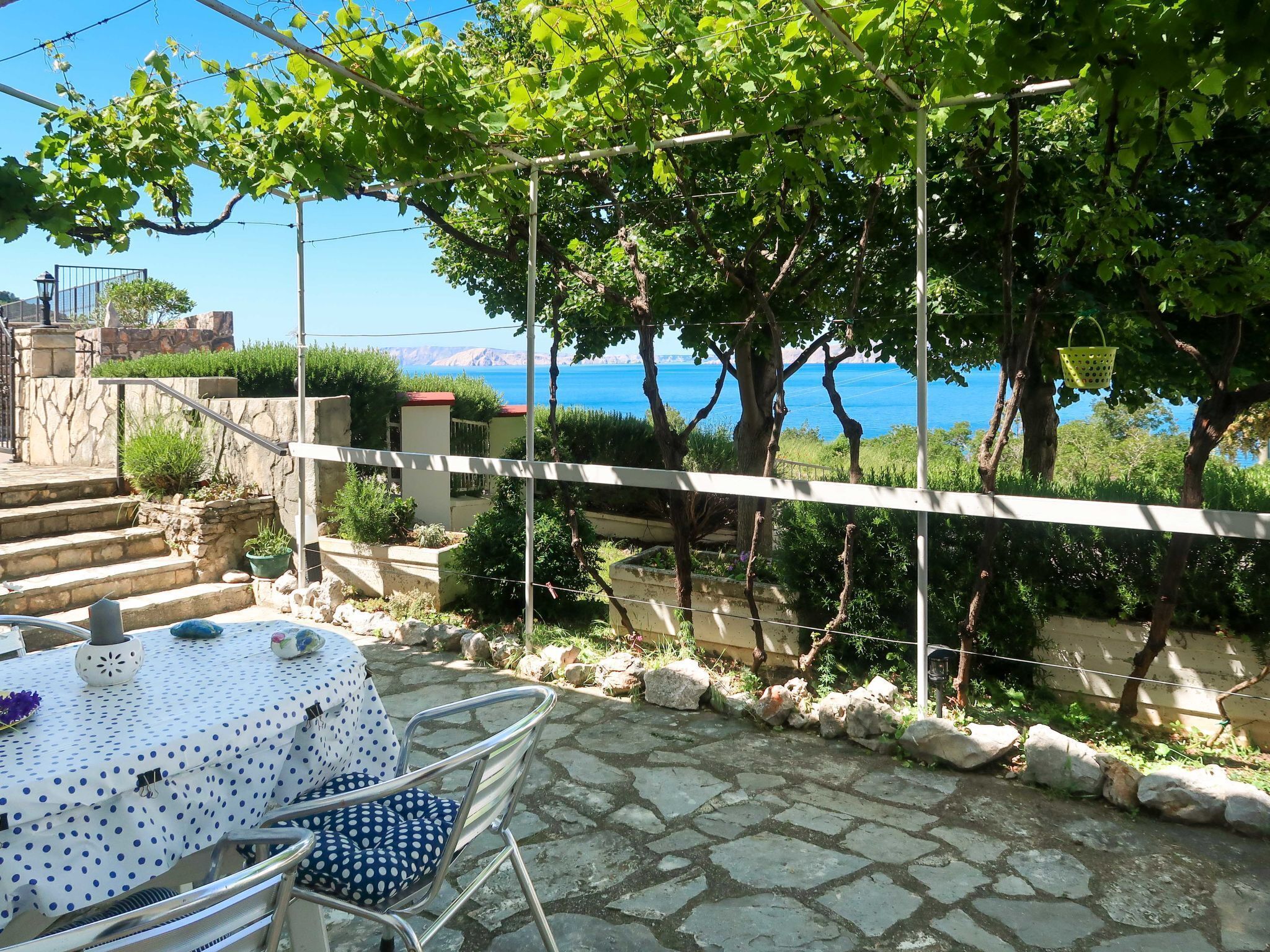 A stone patio with a table and chairs, a view of the sea, and a pergola with vines.