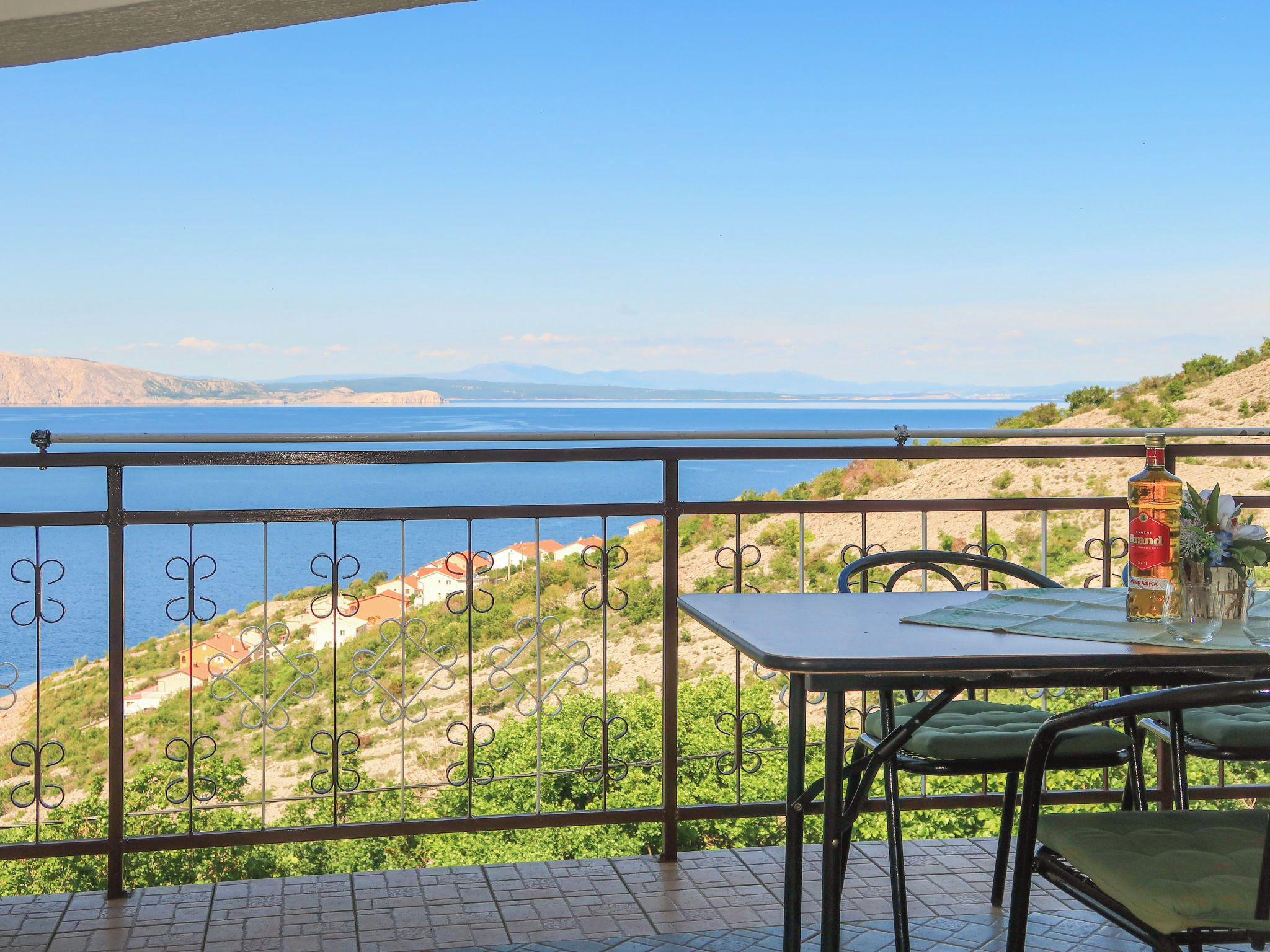 Un balcon avec une table et des chaises, donnant sur un paysage côtier avec des maisons et une étendue d’eau.