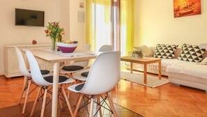 A modern living room with a dining area, featuring a wooden table, white chairs, a sofa with patterned cushions, and a painting on the wall.