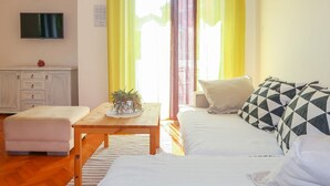 A room with a bed, a sofa, a wooden table with a plant, a TV on the wall, and a window with yellow curtains.