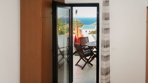 Une pièce dotée d’une porte-fenêtre coulissante donnant sur un balcon extérieur avec une table et des chaises, une vue sur la mer et un parquet à motifs en chevrons.