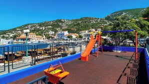 Un parque infantil con tobogán y columpios, con vistas a un puerto deportivo con barcos y un pueblo en la colina al fondo.