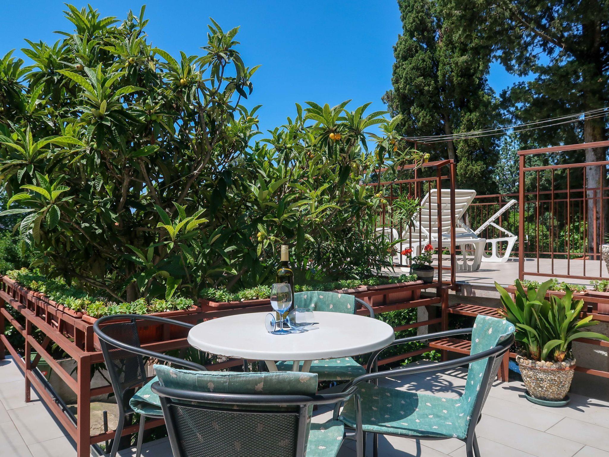 An outdoor patio with a round table and chairs, surrounded by lush greenery and a metal pergola.