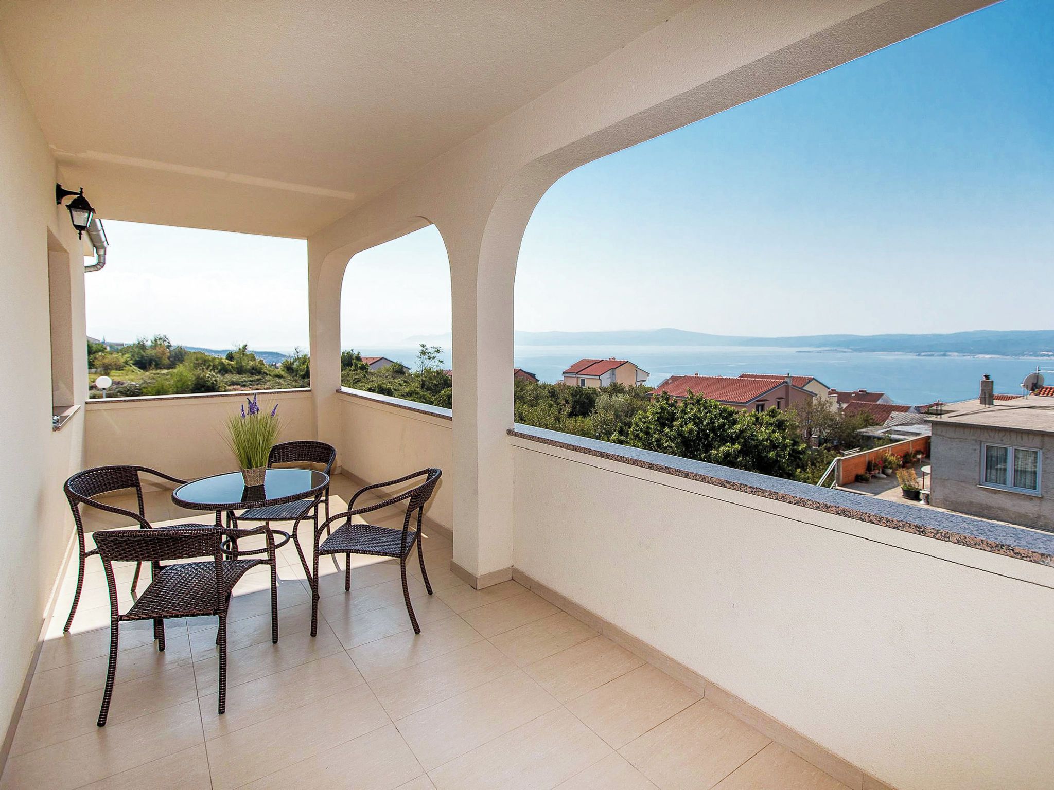 A balcony with a table and chairs, overlooking a coastal landscape with houses and greenery.