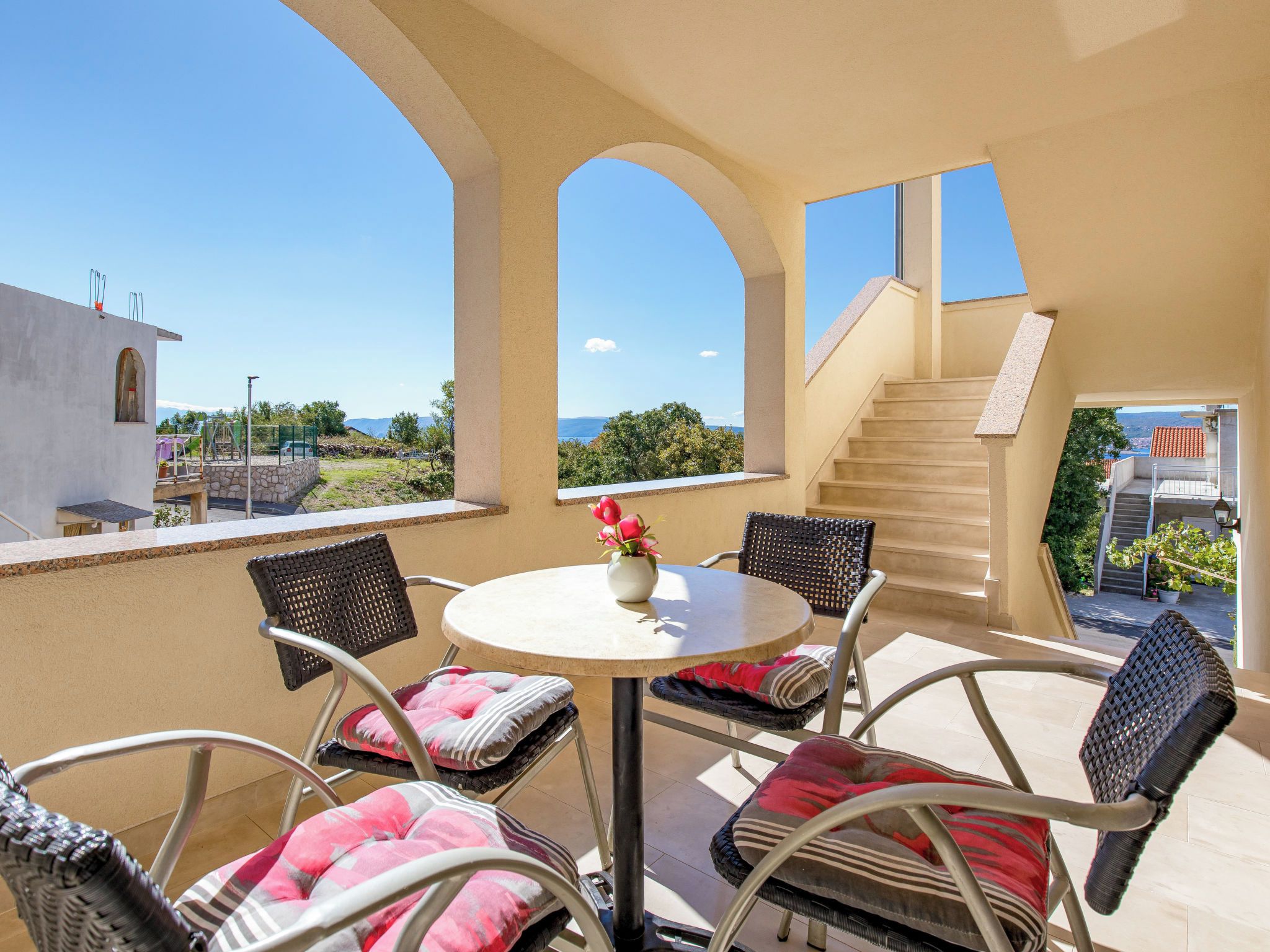 A terrace with a round table and chairs, a flower vase, and a view of the sea and mountains.