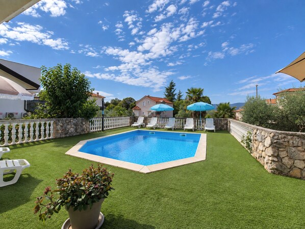 Une piscine rectangulaire bordée de blanc, entourée de gazon synthétique, de chaises longues blanches et d’un mur en pierre.